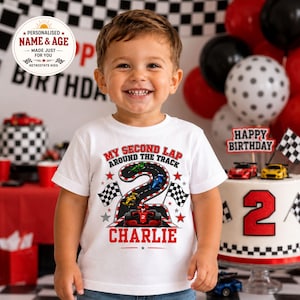 May include: A white short-sleeved t-shirt featuring a graphic of a race track shaped like the number two, with small race cars, checkered flags, and red stars. The text reads My Second Lap Around The Track and the name Charlie in red letters.