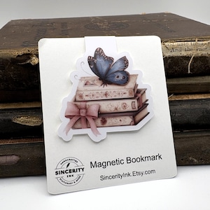 May include: A magnetic bookmark featuring a stack of books with a blue butterfly and a pink bow. The bookmark has the text "Magnetic Bookmark" and "SincerityInk.Etsy.com". The background shows antique books.