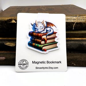 May include: A white magnetic bookmark featuring a sleeping baby dragon atop a stack of books. The dragon is blue and white with orange wings. The books are red, brown, and green. Text on the bookmark reads "Magnetic Bookmark" and "SincerityInk.Etsy.com".