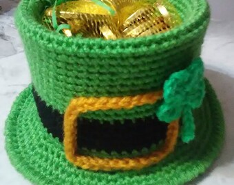 May include: A green crocheted leprechaun hat filled with gold-wrapped chocolate coins. The hat features a black band with a gold buckle and a green shamrock. A festive St. Patrick's Day decoration.