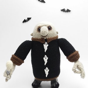May include: A crocheted plush toy of a monster wearing a black coat with brown trim and white ghost buttons. The monster has a large nose and brown eyes. There are three black bats in the background.
