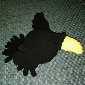 May include: Black plush bird toy with a yellow beak. The bird has a large head and small body. The toy is made of soft, fuzzy fabric.