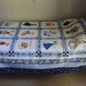 K&ouml;nnte beinhalten: Eine wei&szlig;e und blaue Patchwork-Decke mit gestickten Bildern von holl&auml;ndischen M&auml;dchen, Windm&uuml;hlen und Blumen. Die Decke ist auf einem Holzbettgestell gefaltet.