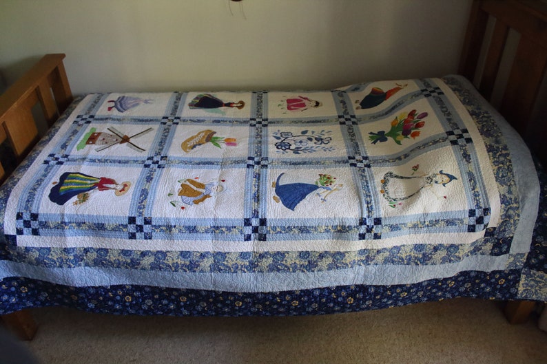 K&ouml;nnte beinhalten: Eine wei&szlig;e und blaue Patchwork-Decke mit gestickten Bildern von holl&auml;ndischen M&auml;dchen, Windm&uuml;hlen und Blumen. Die Decke ist auf einem Holzbettgestell gefaltet.