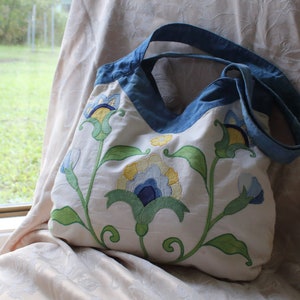 Op de afbeelding: Een witte canvas draagtas met blauwe banden en geborduurde bloemenmotieven in tinten blauw, geel en groen.
