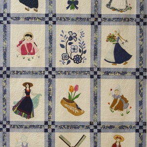 K&ouml;nnte beinhalten: Eine wei&szlig;e Steppdecke mit einem blauen und wei&szlig;en Rand. Die Steppdecke ist in neun Quadrate unterteilt, jedes mit einem anderen gestickten Design. Die Designs beinhalten ein M&auml;dchen in einem blauen Kleid, ein M&auml;dchen in einem roten Kleid, ein M&auml;dchen in einem gelben Kleid, ein M&auml;dchen in einem gr&uuml;nen Kleid, ein M&auml;dchen in einem lila Kleid, ein M&auml;dchen in einem rosa Kleid, ein M&auml;dchen in einem braunen Kleid, ein M&auml;dchen in einem wei&szlig;en Kleid und ein M&auml;dchen in einem blauen Kleid. Die Designs beinhalten auch eine Windm&uuml;hle, ein Paar Holzschuhe, einen Tulpenstrau&szlig; und ein Blumenmuster.
