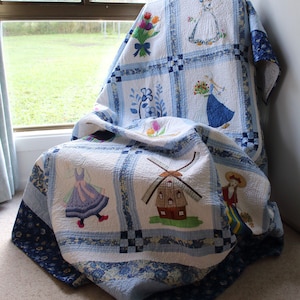 K&ouml;nnte beinhalten: Eine blaue und wei&szlig;e Quiltdecke mit gestickten Bildern von Windm&uuml;hlen, Blumen und Frauen in traditioneller niederl&auml;ndischer Kleidung. Die Decke ist gefaltet und &uuml;ber einen Stuhl gelegt.