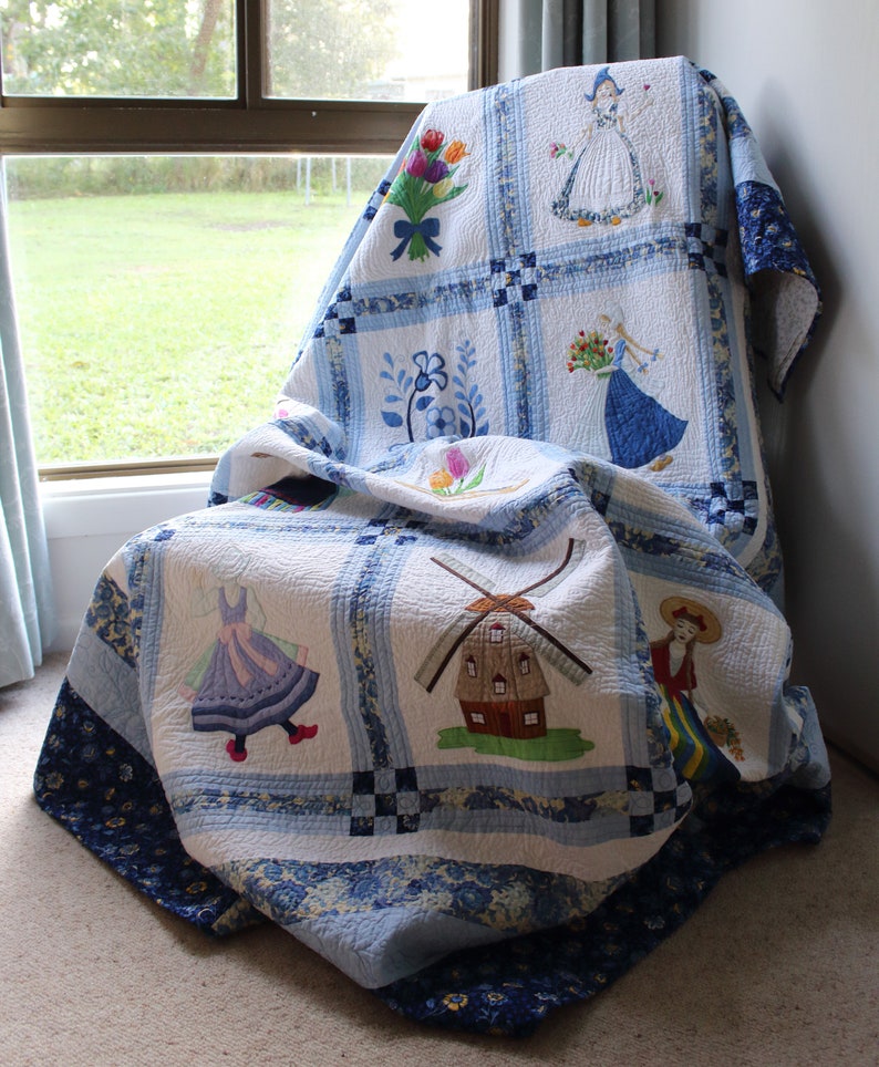 K&ouml;nnte beinhalten: Eine blaue und wei&szlig;e Quiltdecke mit gestickten Bildern von Windm&uuml;hlen, Blumen und Frauen in traditioneller niederl&auml;ndischer Kleidung. Die Decke ist gefaltet und &uuml;ber einen Stuhl gelegt.