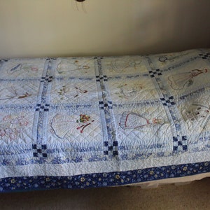 K&ouml;nnte beinhalten: Eine blaue und wei&szlig;e Steppdecke mit gestickten Mustern. Die Steppdecke besteht aus Quadraten mit verschiedenen Mustern, darunter Blumen, V&ouml;gel und andere Motive. Die Steppdecke liegt auf einem Bett mit einer blauen und wei&szlig;en, gemusterten Bettdecke.