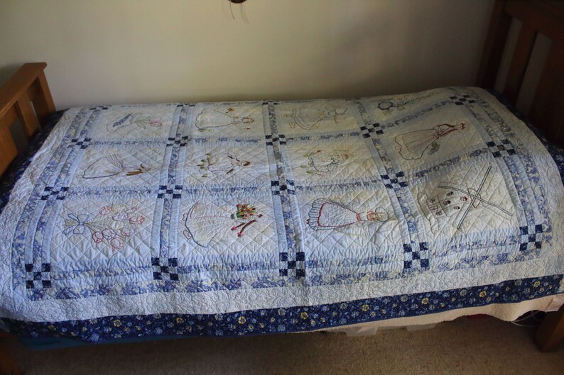 K&ouml;nnte beinhalten: Eine blaue und wei&szlig;e Steppdecke mit gestickten Mustern. Die Steppdecke besteht aus Quadraten mit verschiedenen Mustern, darunter Blumen, V&ouml;gel und andere Motive. Die Steppdecke liegt auf einem Bett mit einer blauen und wei&szlig;en, gemusterten Bettdecke.