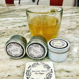 May include: A clear glass of tea sits next to three small metal tins and a coaster. The tins and coaster feature the text "Brooke Birch Tea Shoppe" surrounded by a floral design. The tea is a light amber color.