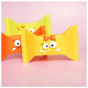 Könnte beinhalten: Drei bunte Papierboxen in Form einer Schleife mit Glitzern und einem Bogen. Die Boxen sind gelb, orange und grün.