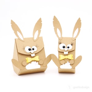 Könnte beinhalten: Zwei braune Papier-Hasen-Geschenkboxen mit Glitzern, weißen Wimpern und gelben Schleifen. Die größere Hasenbox hat eine weiße Wolkenform auf der Vorderseite.