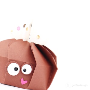 Könnte beinhalten: Eine braune Papierbox mit einem weißen Schmetterling oben drauf. Die Box hat Glitz Augen und ein pinkfarbenes Herz auf der Vorderseite. Der Schmetterling hat goldene und rosafarbene Akzente.