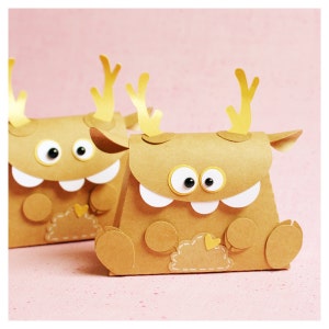 Könnte beinhalten: Zwei braune Papiergeschenkkisten in Form von Monstern mit Glitz Augen, weißen Zähnen und goldenen Geweihen. Die Kisten haben ein gesticktes Herzdetail auf der Vorderseite.