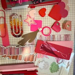 Puede incluir: Una colección de artículos de papelería y accesorios rosas y rojos, que incluyen cuadernos, bolígrafos, marcadores, llaveros y clips. Los artículos presentan motivos de corazón, estrella y libro, con algo de texto visible.