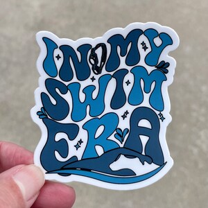 May include: A blue and white sticker with the text "In My Swim Era" in a retro font. The sticker features a silhouette of a swimmer in the water.
