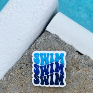 May include: A white sticker with the word "SWIM" repeated three times in blue, light blue, and dark blue lettering. The sticker is on a gray concrete surface.