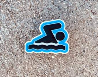 May include: A blue and white swimmer icon with a black silhouette of a person swimming in a pool of water. The icon is made of a rubber-like material.