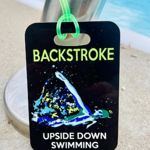 May include: Black rectangular tag with the words "BACKSTROKE" and "UPSIDE DOWN SWIMMING" in white and neon green. The tag features a swimmer illustration in blue, green, and white. A neon green loop is attached to the tag.