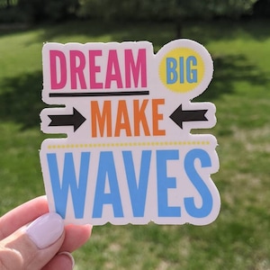 May include: A white sticker with the text "Dream Big Make Waves" in colorful letters. The words "Dream" and "Waves" are in a larger font size than the words "Big" and "Make". The words "Big" and "Make" are surrounded by black arrows pointing in opposite directions.