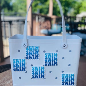 May include: White tote bag with blue "SWIM" stickers. The bag has a white handle and a white base. The stickers are arranged in a pattern on the front of the bag.