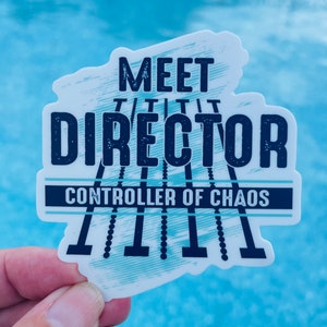May include: A white sticker with blue text that reads "Meet the Director, Controller of Chaos". The text is in a distressed font and is surrounded by a blue and white graphic of a series of lines.