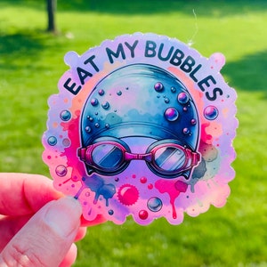 May include: A holographic sticker with a pink, blue, and purple watercolor background. The sticker features a cartoon illustration of a swimmer wearing a blue swim cap and goggles. The text "Eat My Bubbles" is written in a black, bold font around the swimmer.