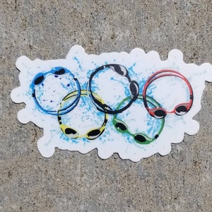May include: A sticker featuring five swimming goggles in the colors blue, yellow, black, green, and red, arranged in the Olympic rings formation. The design includes blue water splash effects on a white background.