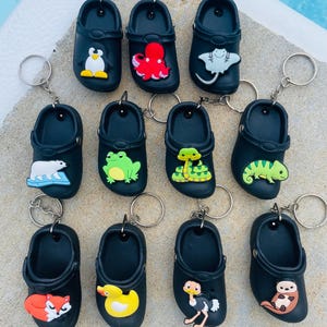 May include: A collection of black clog-shaped keychains, each adorned with a different colorful animal or creature. The charms include a penguin, octopus, stingray, polar bear, frog, snake, chameleon, fox, duck, ostrich, and otter.