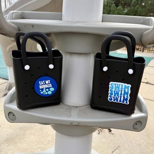 May include: Two black tote bags with handles. One bag has a blue and white circular patch that says "EAT MY BUBBLES". The other bag has a blue and white patch that says "SWIM SWIM SWIM".