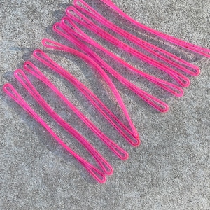 May include: A set of pink hair ties, made of a shiny, flexible material. The hair ties are arranged in a criss-cross pattern on a gray surface.