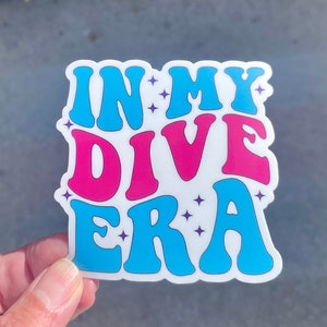 May include: A sticker with the text "In My Dive Era" in a retro font. The words "In" and "Era" are blue, and the word "Dive" is pink. There are small purple stars around the text.