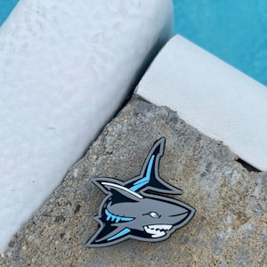 May include: A gray enamel pin of a shark with blue accents. The shark is facing right and has a toothy grin. The pin is on a gray concrete surface.