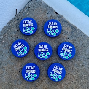 May include: Seven blue circular charms with the text "EAT MY BUBBLES" in white, surrounded by teal bubbles. The charms are arranged on a gray concrete surface, with a blue water background.