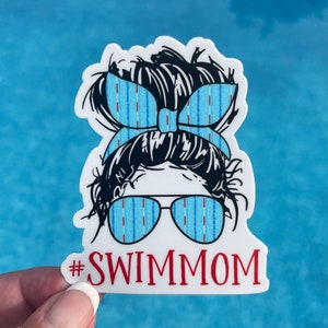 May include: White sticker with a cartoon illustration of a woman with a messy bun, wearing blue and red striped sunglasses and a blue and red striped headband. The text "#SWIMMOM" is below the illustration.