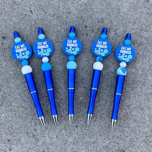 May include: Five blue pens with blue and white bead accents and a blue round charm with the text "Eat My Bubbles" on each pen.