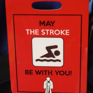 May include: Red luggage tag with white text that reads "May the stroke be with you!"  A white silhouette of a person swimming is in the center of the tag. A white silhouette of a person wearing a robe is at the bottom of the tag.