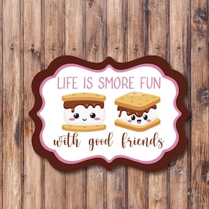 May include: A white sign with brown and pink borders features the text "Life is s'more fun with good friends". Two cartoon s'mores with smiling faces are depicted on the sign.
