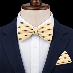 May include: A cream-colored bow tie and matching pocket square featuring a repeating pattern of small, detailed bumblebees with yellow and black bodies. The set is displayed on a white collared shirt worn under a dark navy blue suit jacket.