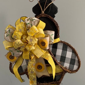 May include: A black and white checkered bee-shaped wreath with a yellow and brown bow decorated with sunflowers and honeycomb patterns.