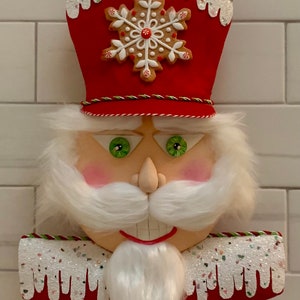 Puede incluir: Un adorno de cascanueces navideño rojo y blanco con un copo de nieve de jengibre en el sombrero. El cascanueces tiene barba y bigote blancos, ojos verdes y boca roja.