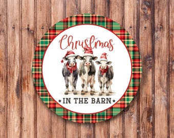 May include: A round Christmas-themed sign with a red and green plaid border. The center features a white background with the text "Christmas in the barn" and three cows wearing Santa hats.