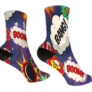 May include: A pair of comic book-themed socks. The socks feature a blue background with white speech bubbles containing words like "BOOM!" and "BANG!" in bold, red and black lettering. The design includes yellow and orange explosions and black accents.