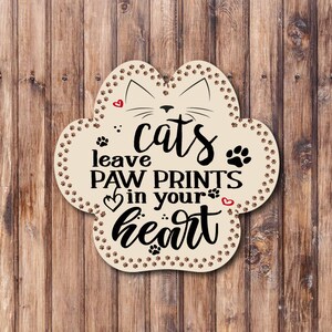 Könnte beinhalten: Ein pfotenförmiges Holzschild mit dem Text "cats leave paw prints in your heart". Das Schild hat ein Katzengesicht-Design und Pfotenabdrücke an den Rändern. Rote Herzen sind ebenfalls enthalten.