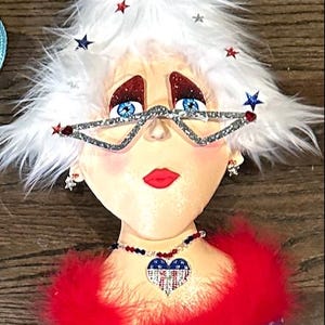 Puede incluir: Una cabeza de muñeca blanca y esponjosa con un boa de plumas rojo y un collar en forma de corazón azul y blanco con estrellas. La muñeca lleva gafas brillantes y tiene estrellas rojas y azules en su cabello blanco.