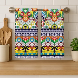 May include: Two decorative kitchen towels with a vibrant floral design. The towels feature a colorful pattern with red, orange, yellow, blue, and green elements. They are hanging on a metal bar against a white tiled wall.