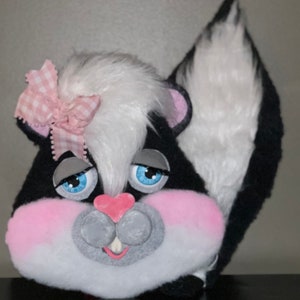 May include: A plush black and white skunk toy with a pink bow, blue eyes, and a pink nose. The toy has a white fluffy tail and a black body.