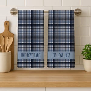 May include: Two blue plaid kitchen towels hanging on a silver bar. Each towel has a light blue band with the text "LIVE LOVE LAKE" in dark blue. The towels are displayed on a wooden countertop with a white tile backsplash.