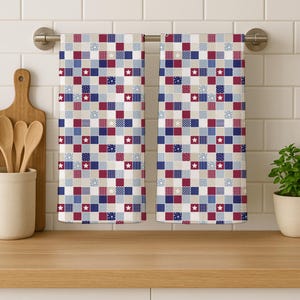 May include: Two kitchen towels with a patchwork design. The towels feature a pattern of squares in red, white, blue, and beige, with star and polka dot accents. They are hanging on a silver towel rack above a wooden countertop.